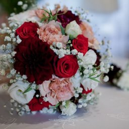 Pro SmArt Studio Adam Kowal - Bukiet ślubny z ciemnoczerwonych dalii, czerwonych róż, białych eustom i bladoróżowych goździków, otoczony drobnymi białymi kwiatami gipsówki na białym obrusie.
