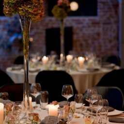 Pro SmArt Studio Adam Kowal - Elegancko nakryty stół weselny z wysokimi wazonami z różowymi i zielonymi kwiatami, świecami, szklankami i szarymi serwetkami z subtelnym wzorem ryb na tle ceglanej ściany.