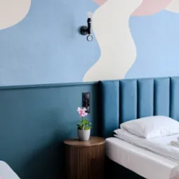 Pok&oacute;j hotelowy z ł&oacute;żkiem, stolikiem nocnym z kwiatem w doniczce, lampą w stylu industrialnym i abstrakcyjnym malowaniem na ścianie w pastelowych kolorach.