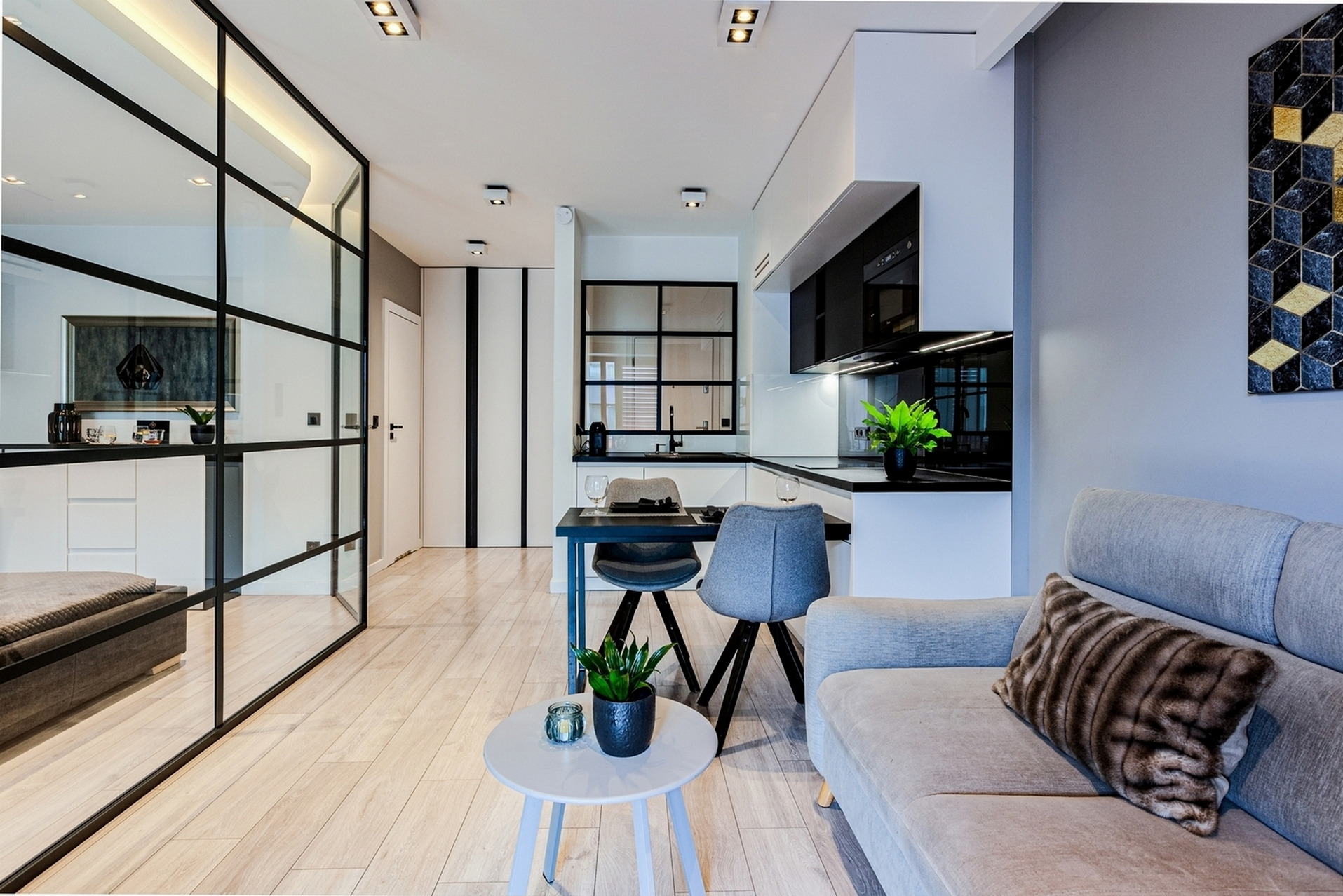 Nowoczesne wnętrze mieszkania z przeszkloną ścianą działową, drewnianą podłogą, szarą sofą z wzorzystą poduszką i czarno-białą kuchnią w tle.