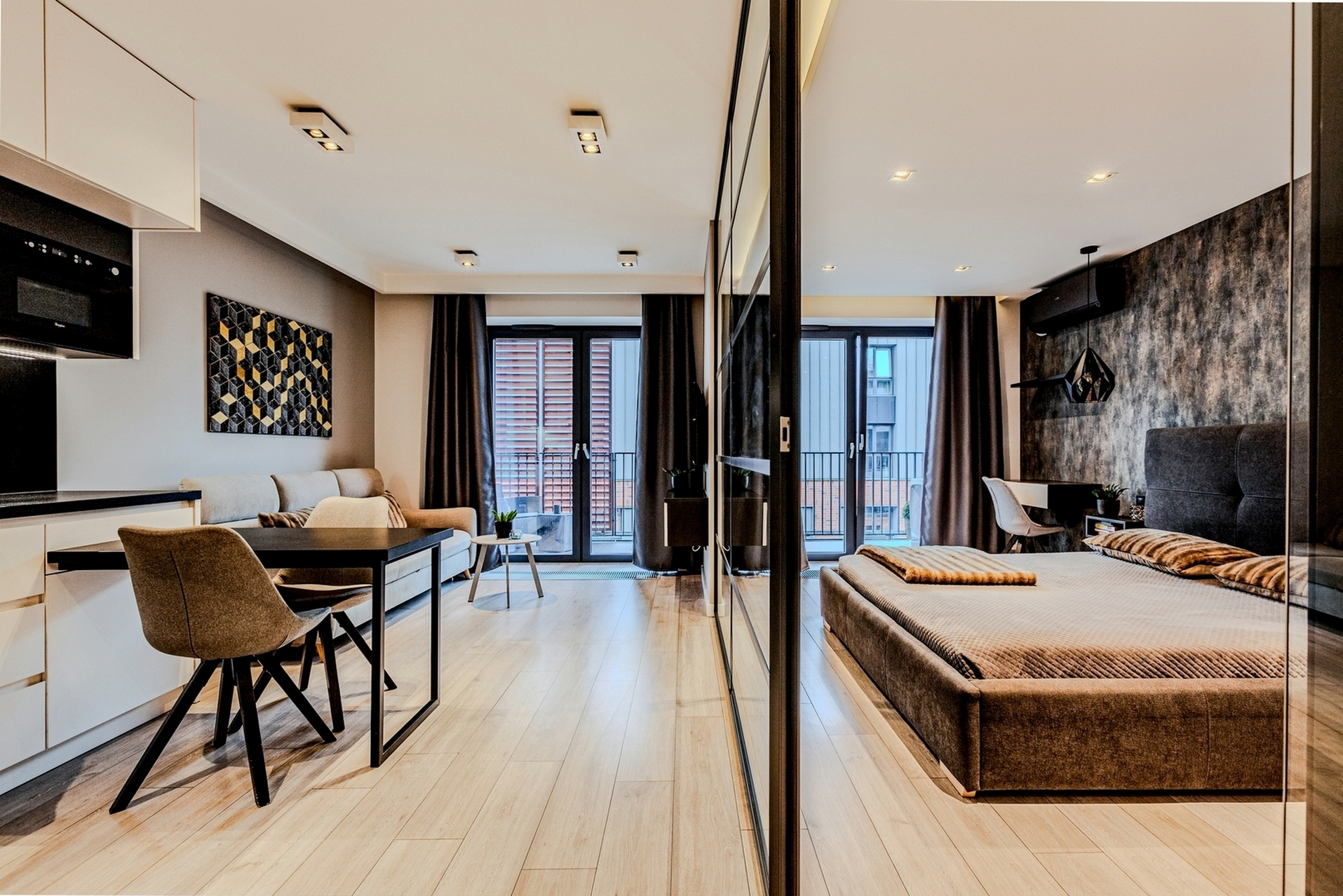 Przestronne studio apartament z otwartą kuchnią, jadalnią i sypialnią, oddzielone szklaną ścianą; widok na balkon z obu części mieszkania.