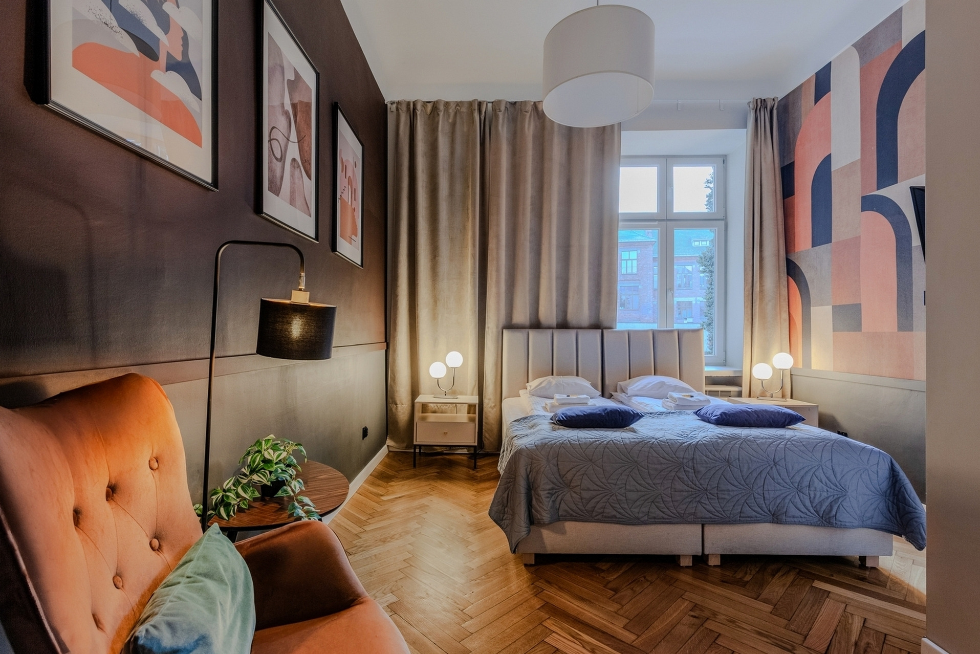 Przytulna sypialnia z parkietem w jodełkę, łóżkiem z szarym zagłówkiem i kołdrą, pomarańczowym fotelem z zieloną poduszką, designerskimi lampkami nocnymi i abstrakcyjnymi obrazami na ścianach...