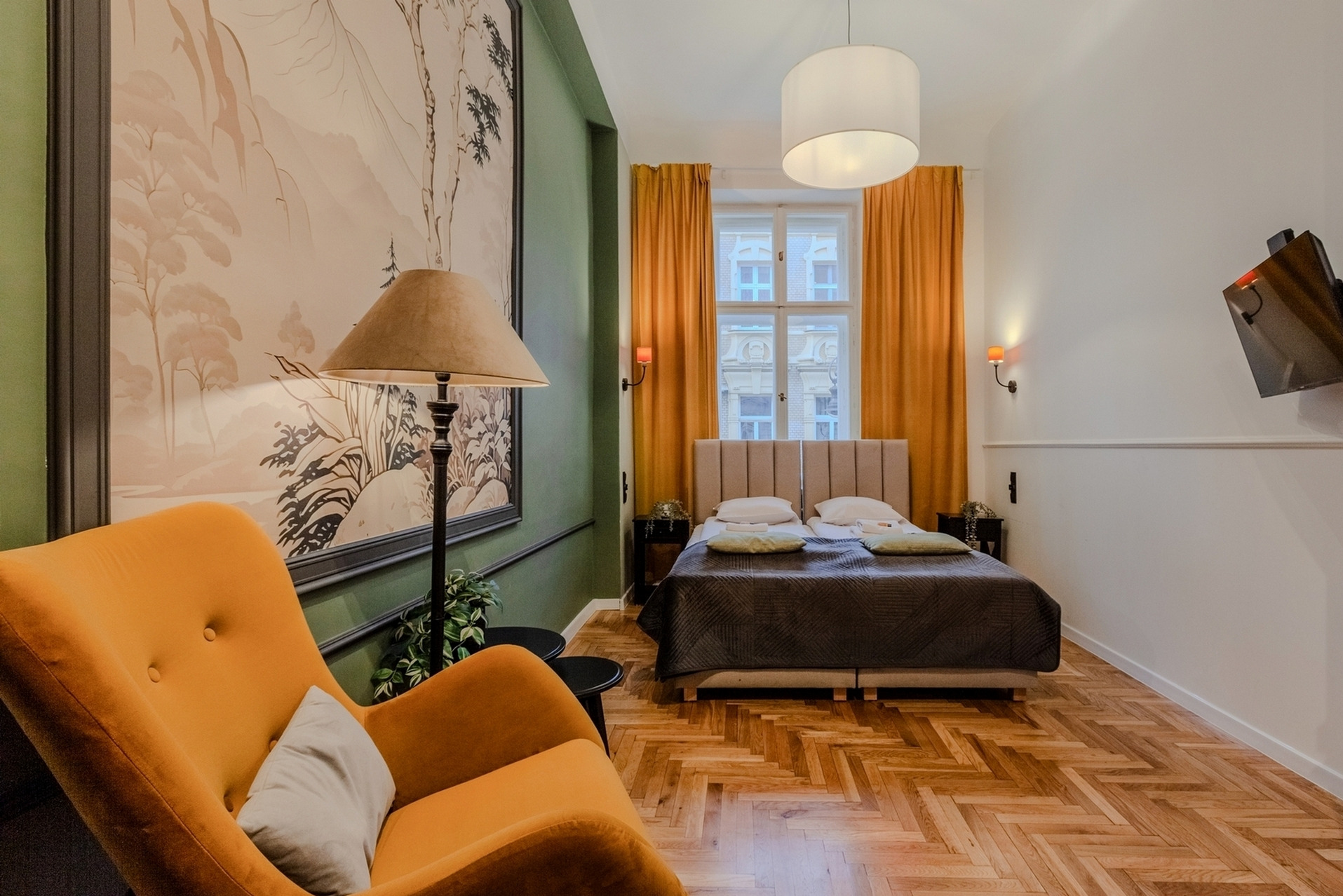 Pokój hotelowy z podwójnym łóżkiem, pomarańczowym fotelem, parkietem w jodełkę, obrazem na ścianie w odcieniach beżu i zieleni oraz pomarańczowymi zasłonami.