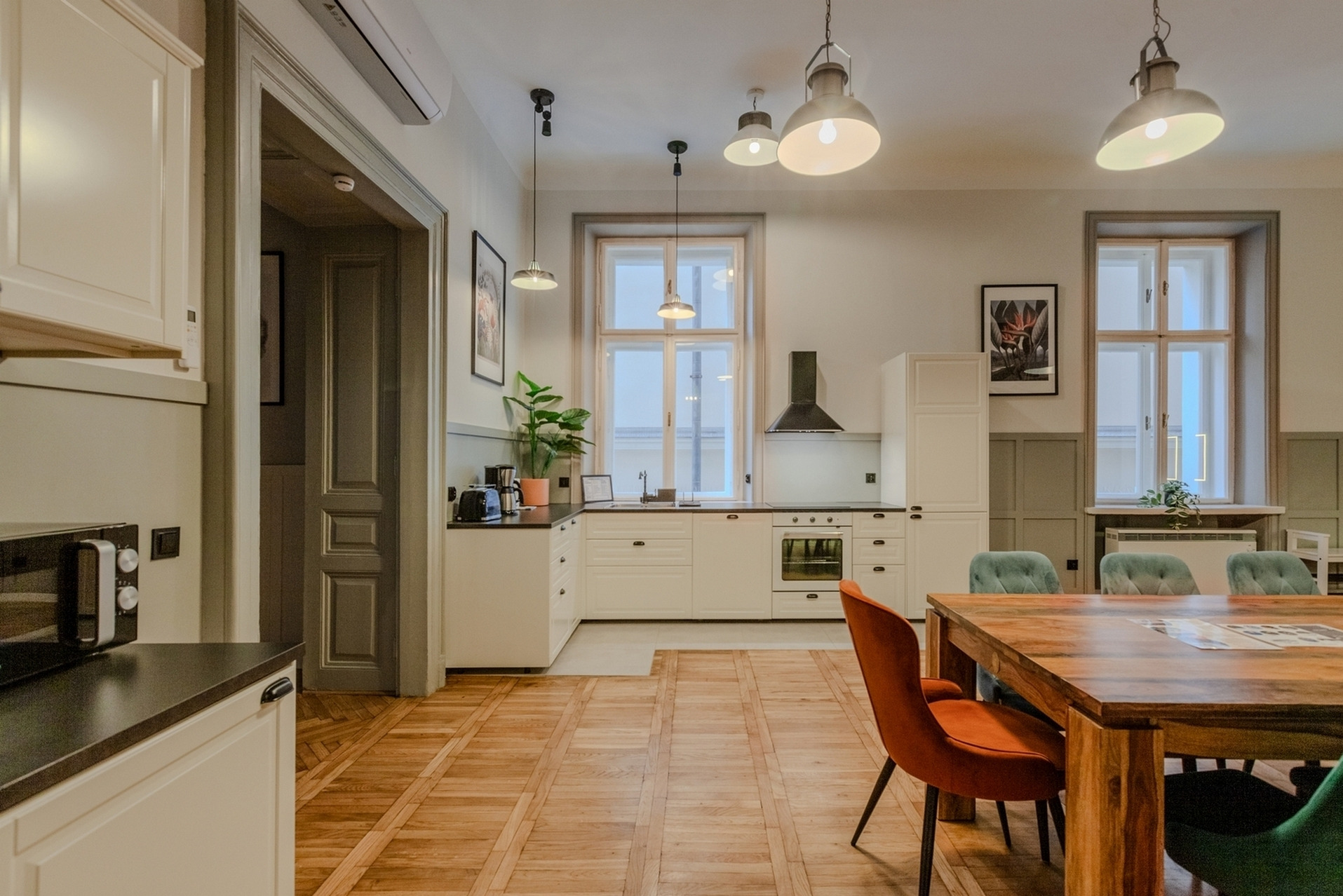 Jasna kuchnia z drewnianą podłogą w jodełkę, pomarańczowe i zielone krzesła przy drewnianym stole, okna z widokiem na zewnątrz, nowoczesne oświetlenie wiszące.