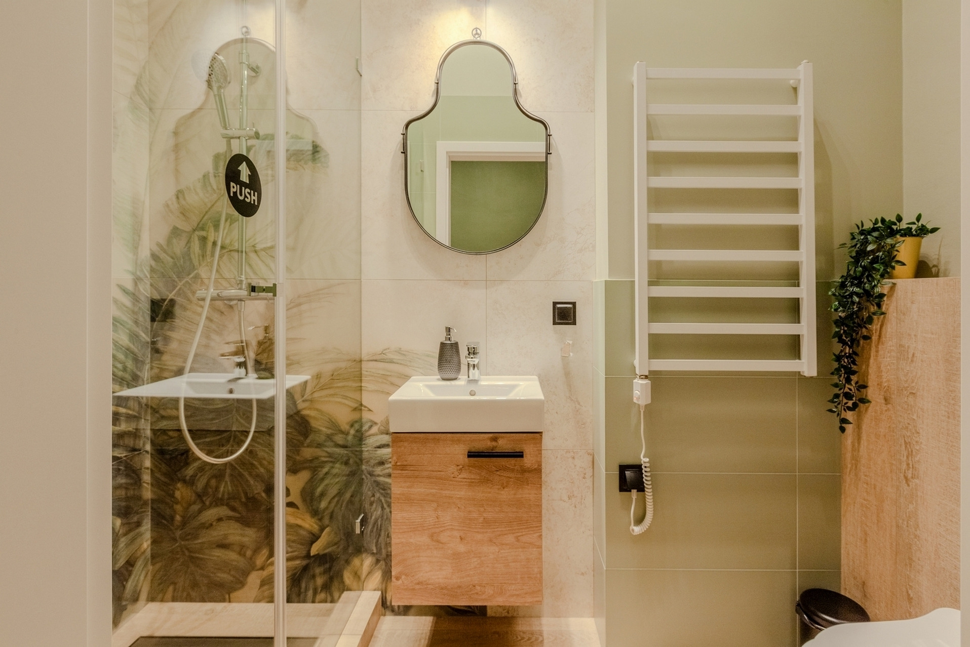 Nowoczesna łazienka z kabiną prysznicową ze szklanymi drzwiami z naklejką 'PUSH', umywalką na drewnianej szafce, ozdobnym lustrem i grzejnikiem drabinkowym z rośliną w żółtej doniczce.