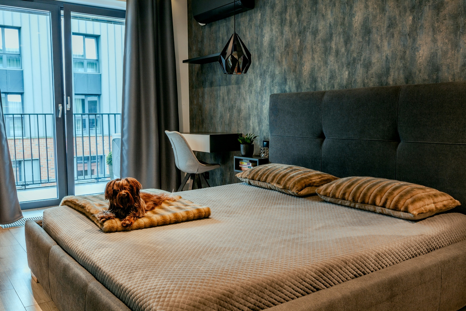 Nowoczesna sypialnia z łóżkiem z szarą tapicerką, dekoracyjnymi poduszkami w odcieniach brązu i beżu, oraz małym psem leżącym na pledzie; w tle biurko z krzesłem, balkon z widokiem na budynek