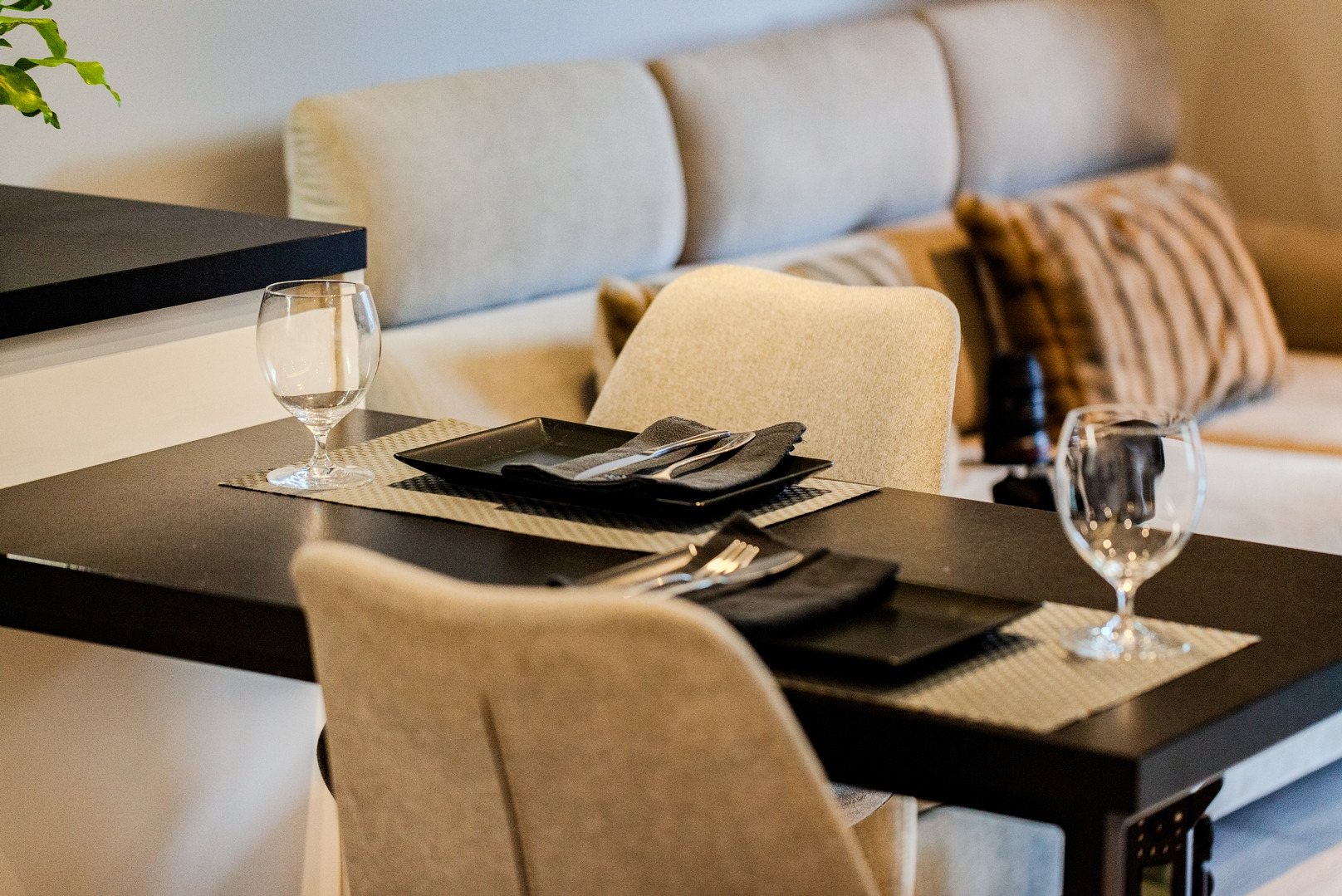 Stół z czarnym blatem, nakryty dla dwóch osób, z czarnymi kwadratowymi talerzami, sztućcami i szarymi serwetkami, ustawiony w nowoczesnym wnętrzu z beżową sofą w tle.