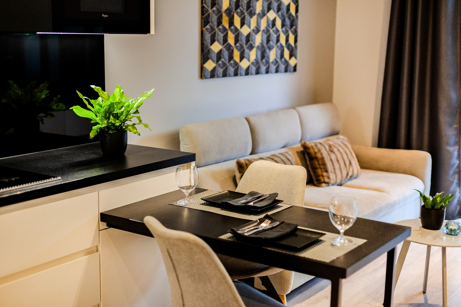 Nowoczesne wnętrze: stół z czarnymi talerzami i sztućcami, szklanki, beżowa sofa z poduszkami, roślina w czarnej doniczce, geometryczny obraz na ścianie.