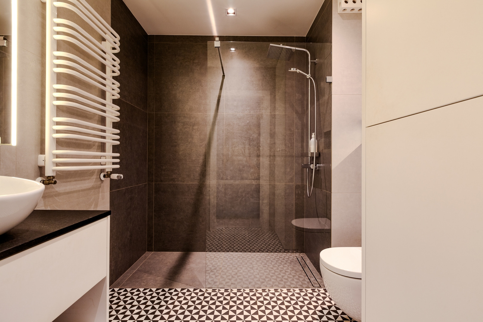 Nowoczesna łazienka z szarą kabiną prysznicową, geometryczną posadzką, grzejnikiem drabinkowym i umywalką nablatową na ciemnym blacie.