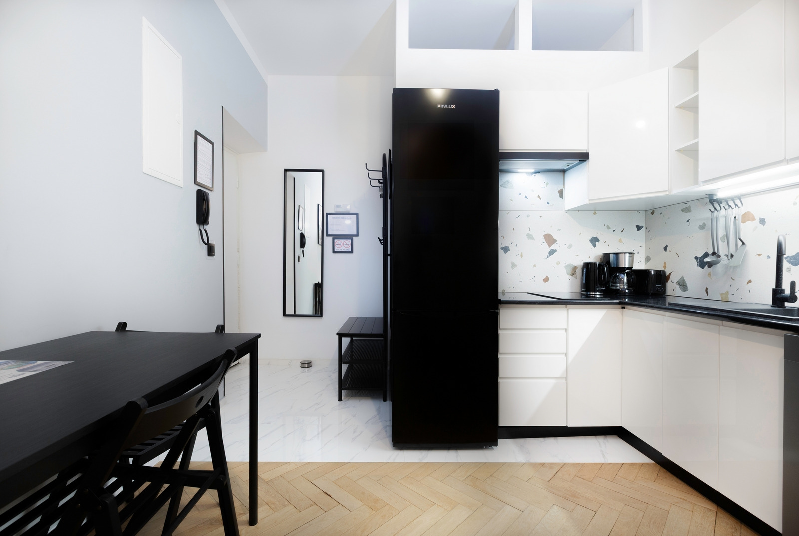 Nowoczesna kuchnia z białymi szafkami, czarnym blatem i lodówką, oraz mozaikową ścianą nad blatem. W tle stół z krzesłami i lustro w czarnej ramie.