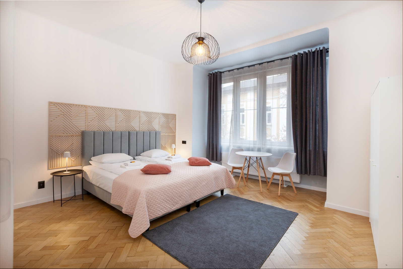 Jasne wnętrze sypialni z drewnianą podłogą w jodełkę, łóżkiem z szarym zagłówkiem i geometrycznym panelem, okrągłym stolikiem z krzesłami przy oknie oraz nowoczesną lampą wiszącą.