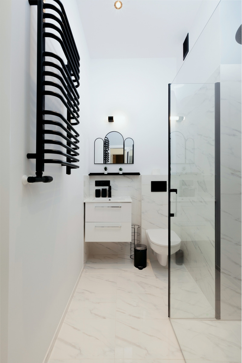 Minimalistyczna łazienka w bieli i czerni z grzejnikiem drabinkowym, trzema lustrami nad umywalką i kabiną prysznicową z marmurowym wykończeniem.