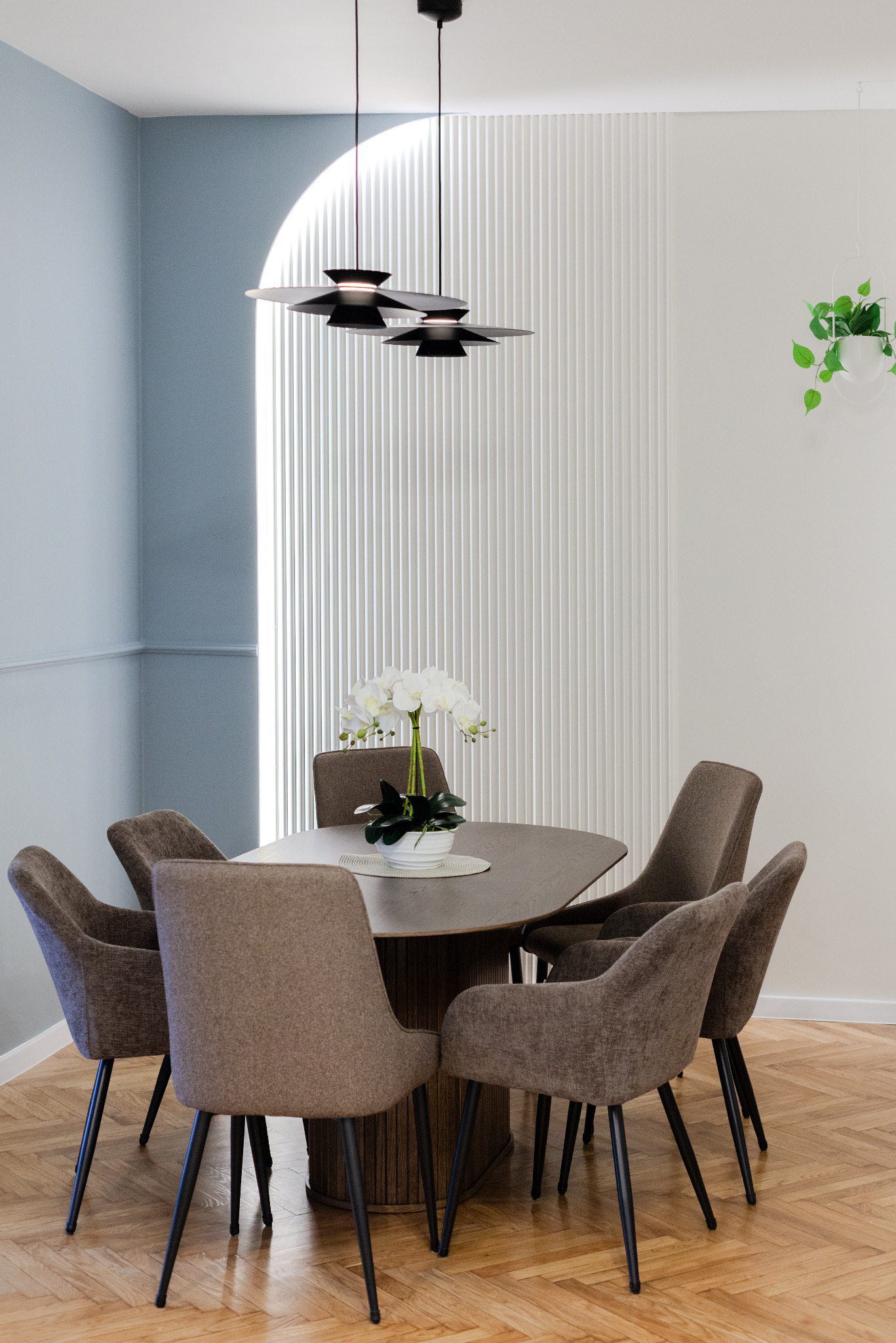 Nowoczesna jadalnia z owalnym stołem, sześcioma tapicerowanymi krzesłami na czarnych nogach i designerską lampą wiszącą z trzema kloszami, oświetlona światłem wpadającym przez okno z dekoracyjną...