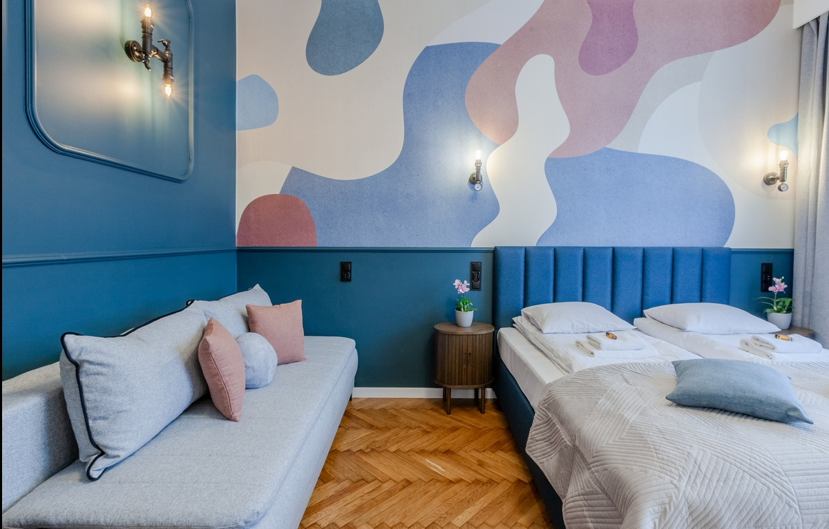 Nowoczesny pokój hotelowy z dwoma łóżkami, sofą, drewnianą podłogą w jodełkę i abstrakcyjną tapetą w pastelowych kolorach.