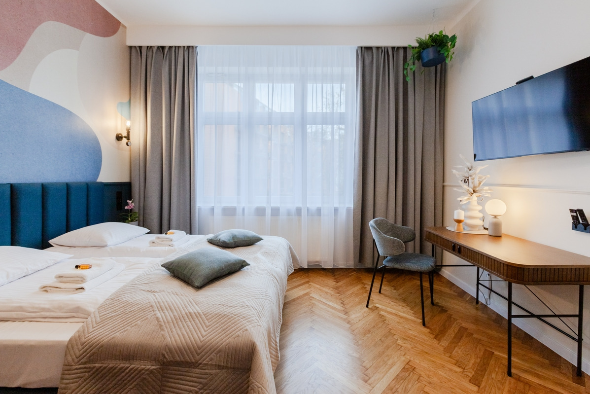 Nowoczesna sypialnia z dwoma łóżkami, parkietem w jodełkę, biurkiem z dekoracjami i telewizorem na ścianie, oświetlona naturalnym światłem.