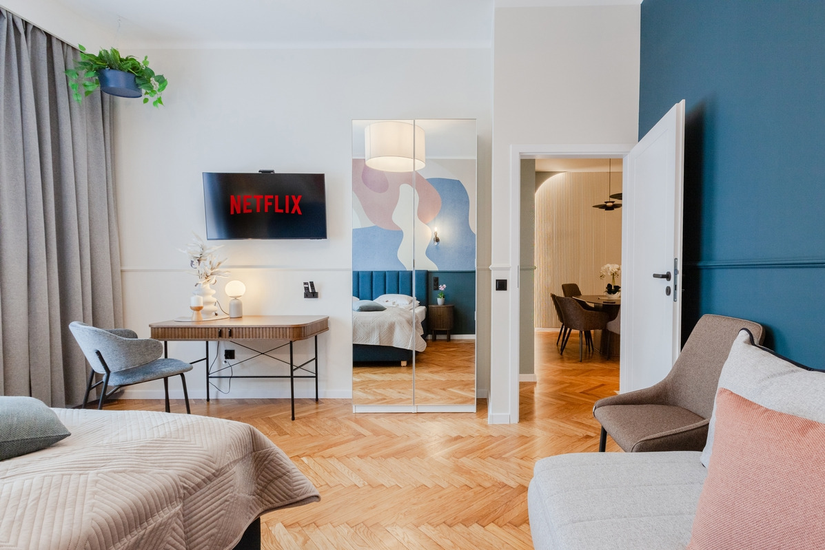 Jasne wnętrze apartamentu z parkietem w jodełkę, biurkiem pod telewizorem z logo Netflix i lustrem odbijającym sypialnię, widok na jadalnię w tle.