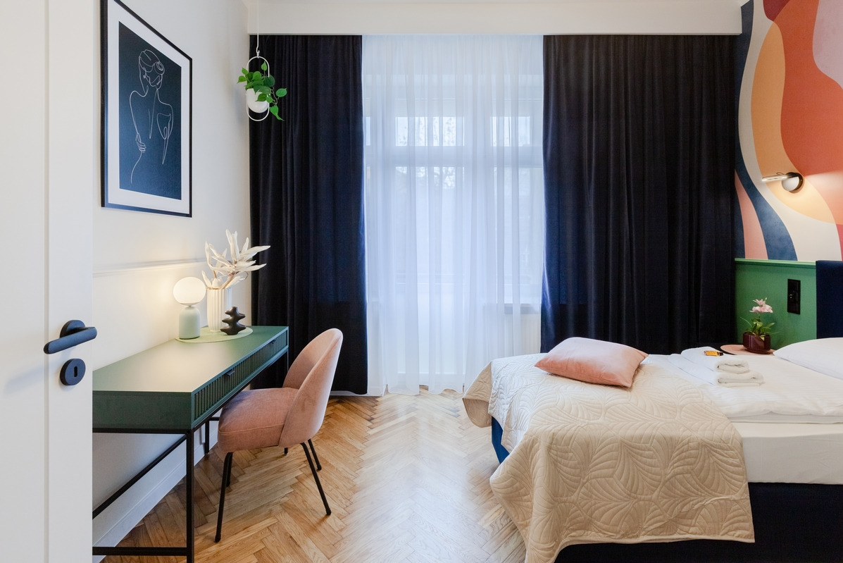 Pokój hotelowy z łóżkiem, biurkiem w kolorze butelkowej zieleni, pastelowym krzesłem i parkietem w jodełkę. Nad biurkiem grafika w czarnej ramie i wisząca roślina. Ściana za łóżkiem z abstrakcyjnym...