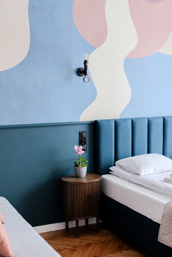 Pokój hotelowy z łóżkiem, stolikiem nocnym z kwiatem w doniczce, lampą w stylu industrialnym i abstrakcyjnym malowaniem na ścianie w pastelowych kolorach.