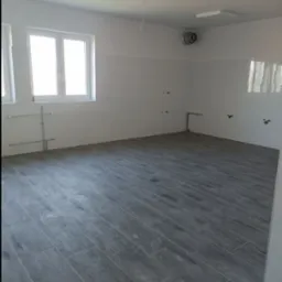 Puste pomieszczenie po remoncie z szarymi płytkami imitującymi drewno na podłodze, białymi płytkami na ścianach, widocznymi instalacjami hydraulicznymi i wentylacją sufitową.