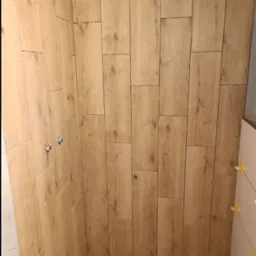 Ściana łazienki w trakcie układania płytek imitujących drewno, widoczne krzyżyki dystansowe, wystające rury instalacji wodnej oraz karton po płytkach na podłodze.