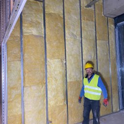 Uslugi remontowe - Pracownik w żółtym kasku i kamizelce odblaskowej stoi na tle ściany z metalowym stelażem wypełnionym wełną mineralną, widoczne instalacje pod sufitem.