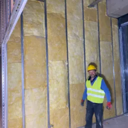 Pracownik w żółtym kasku i kamizelce odblaskowej stoi na tle ściany z metalowym stelażem wypełnionym wełną mineralną, widoczne instalacje pod sufitem.