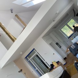 Uslugi remontowe - Wnętrze poddasza w trakcie prac wykończeniowych: widoczne drewniane belki konstrukcyjne, okno dachowe, białe ściany, okno balkonowe zabezpieczone taśmą malarską, oraz elementy wyposażenia w trakcie...