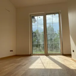 Jasne, puste pomieszczenie z drewnianą podłogą, dużym oknem z widokiem na zieleń i wystającymi kablami ze ściany, wpadające promienie słoneczne.