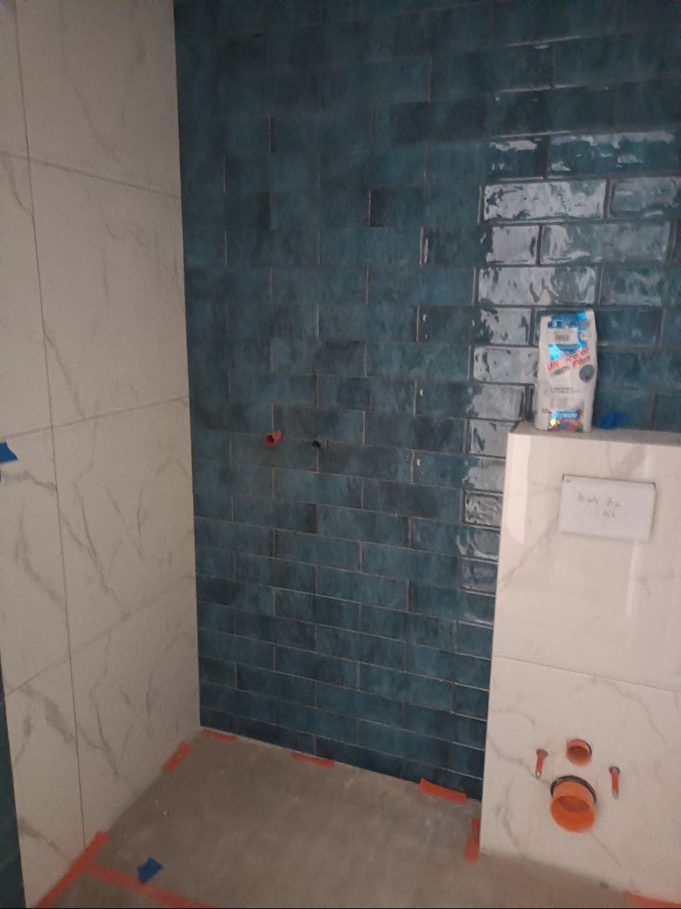 Błękitne, błyszczące płytki ułożone na ścianie łazienki w trakcie prac wykończeniowych, widoczny worek z zaprawą do fugowania i fragment instalacji podtynkowych.