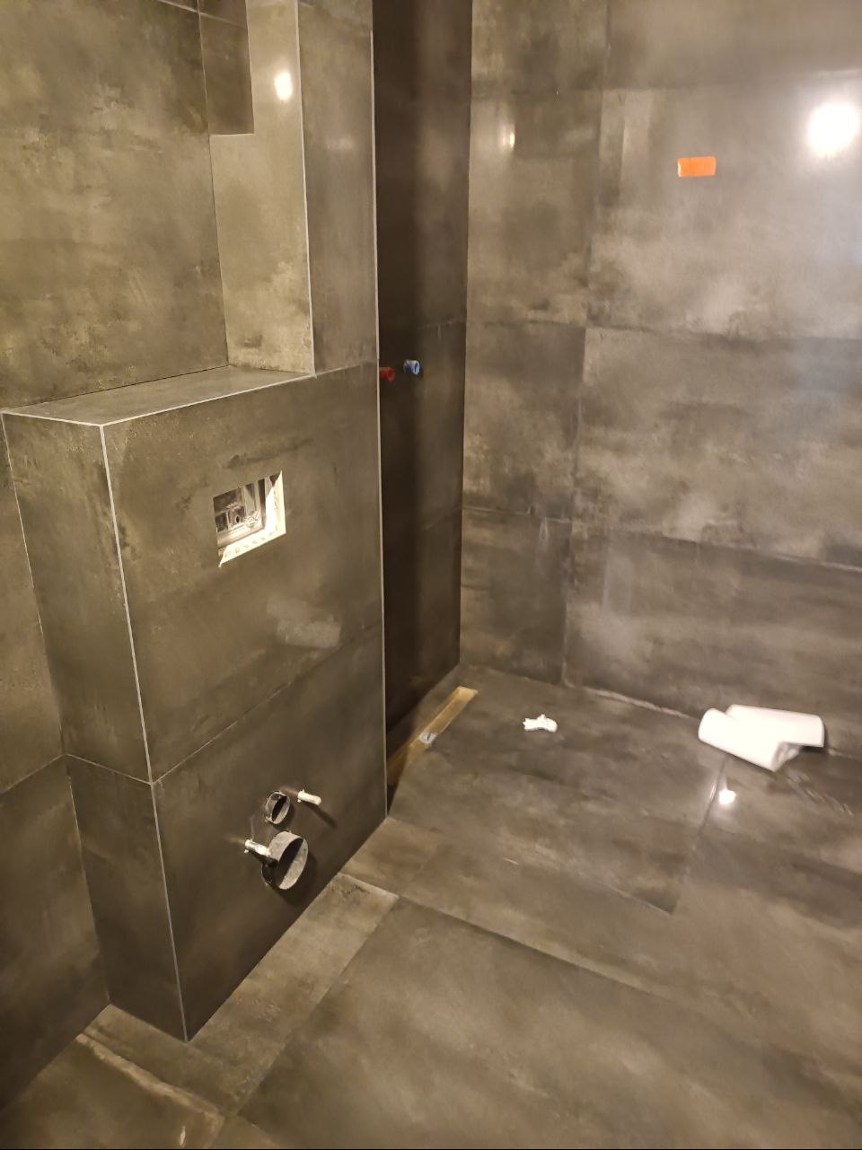 Szara łazienka w trakcie wykończenia: widoczne przyłącza hydrauliczne, zamontowana podtynkowa spłuczka WC oraz ręczniki na podłodze