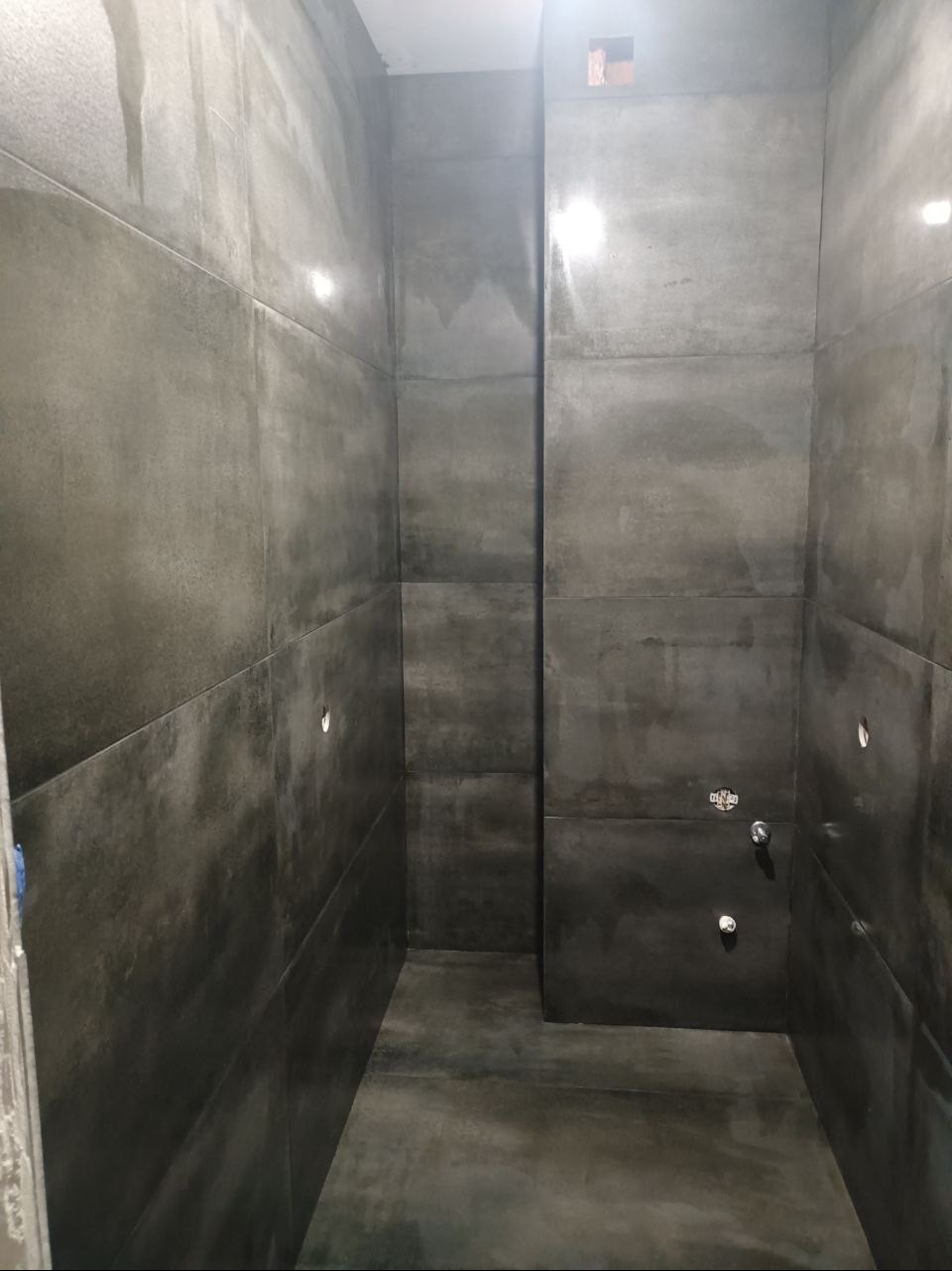 Surowa, ciemnoszara łazienka w trakcie wykończenia, z dużymi płytami imitującymi beton na ścianach i podłodze, widoczne otwory na armaturę i brakujące elementy wykończeniowe.