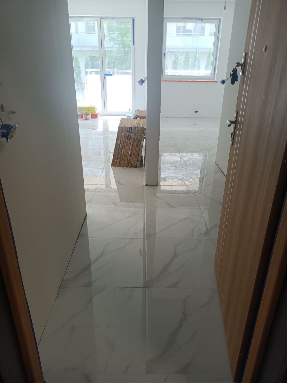 Ułożone białe płytki marmurowe na podłodze w trakcie prac wykończeniowych, widoczne materiały budowlane i otwarte drzwi wejściowe.