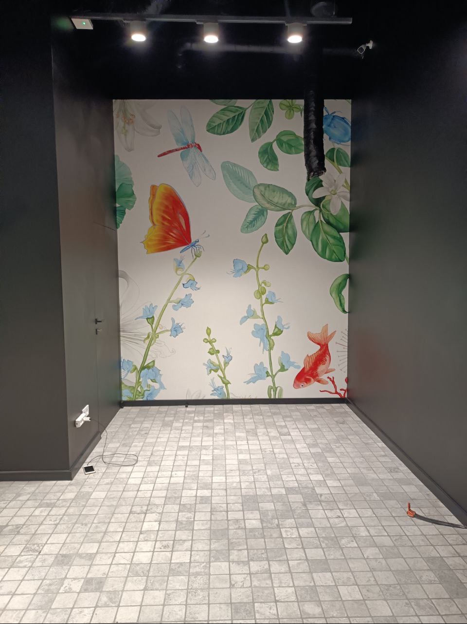 Pusta przestrzeń z szarą, kwadratową podłogą wyłożoną płytkami, ściany pomalowane na ciemnoszaro, na jednej ścianie fototapeta z motywem akwarelowych kwiatów, motyli i ryb.