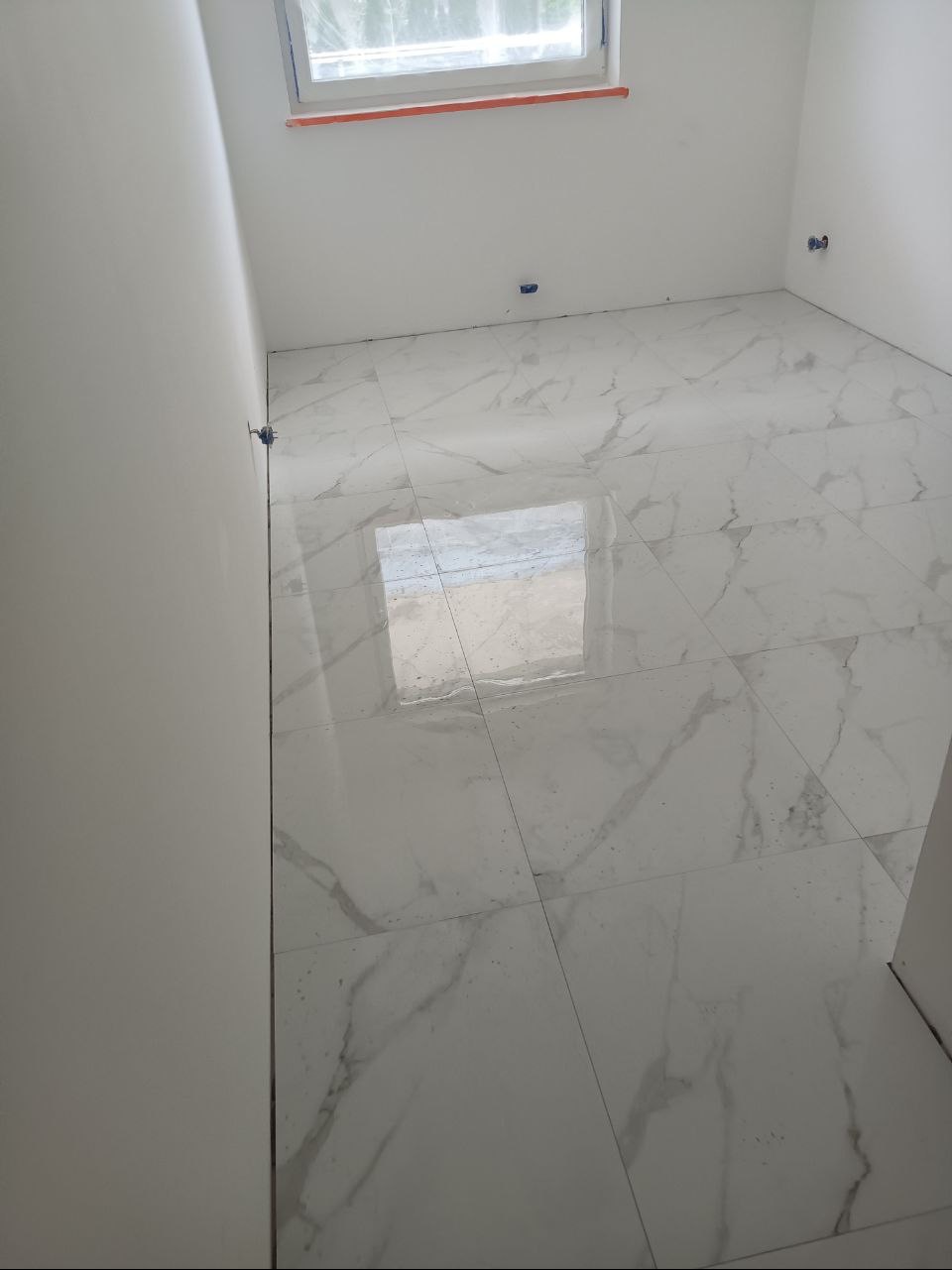 Ułożone białe płytki podłogowe z szarym marmurkowym wzorem w pustym pokoju z widocznym oknem i wystającymi przewodami elektrycznymi ze ściany.