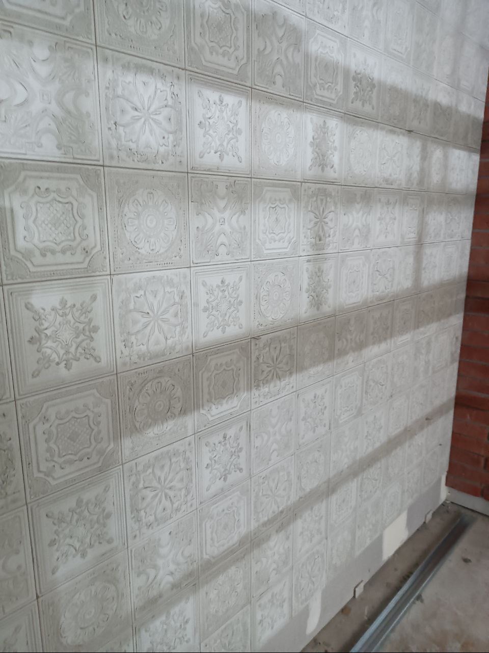 Ściana wyłożona kwadratowymi, jasnoszarymi płytkami z wytłaczanymi wzorami kwiatowymi i geometrycznymi, widoczne niedociągnięcia w fugowaniu, ujęcie z boku z widocznym fragmentem ceglanej ściany...