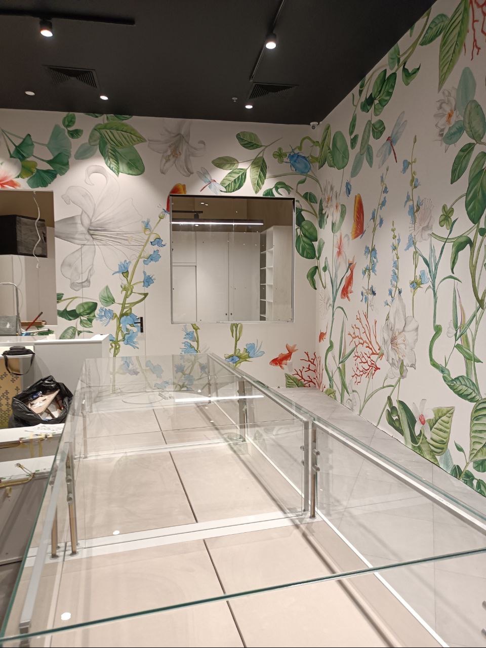Wnętrze komercyjne w trakcie wykańczania: ściany z motywem akwarelowych kwiatów i ryb, szklane witryny sklepowe w trakcie montażu, lustro w ramie na ścianie.