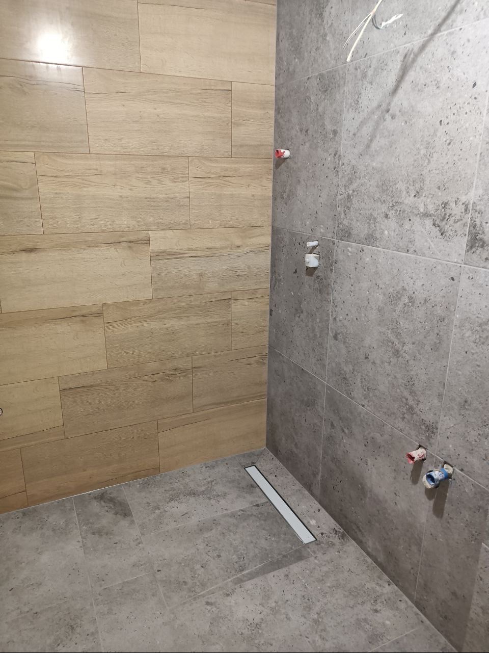 Fragment łazienki w trakcie remontu: Ściana wyłożona płytkami imitującymi drewno sąsiaduje z szarą ścianą z płytek o nieregularnej fakturze. Widoczne niezamaskowane rury i odpływ liniowy w podłodze.