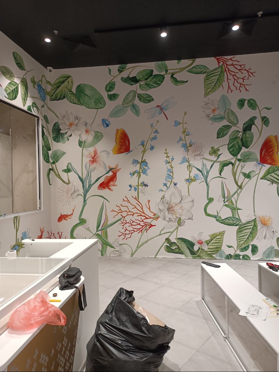 Ściana w trakcie dekorowania z motywem akwarelowych kwiatów, ryb i owadów, widoczne niedoskonałości malowania i śmieci budowlane na podłodze.