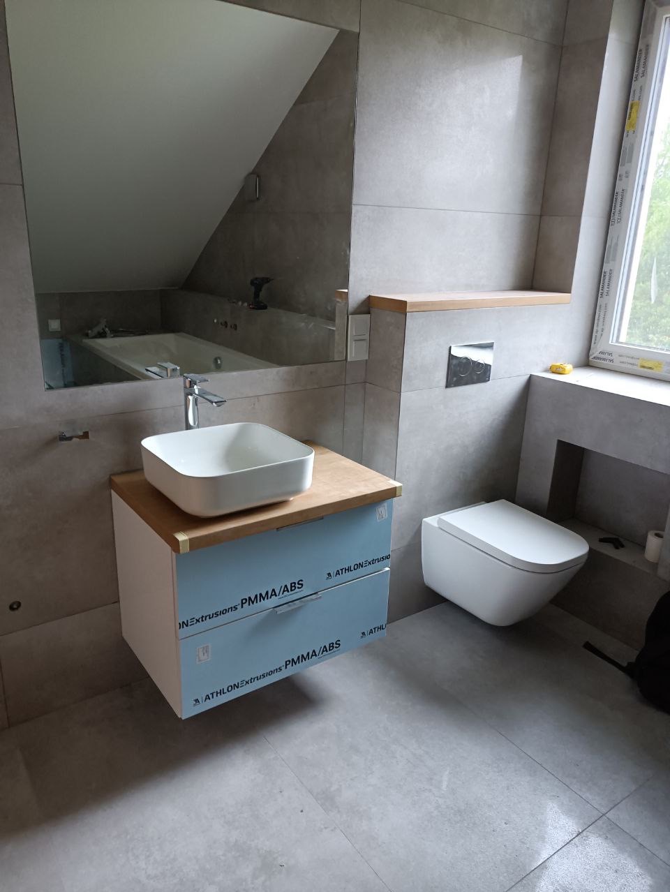 Nowoczesna łazienka w szarościach z podwieszaną szafką umywalkową z blatem drewnopodobnym, wiszącą toaletą i dużym lustrem odbijającym wannę pod skosem.