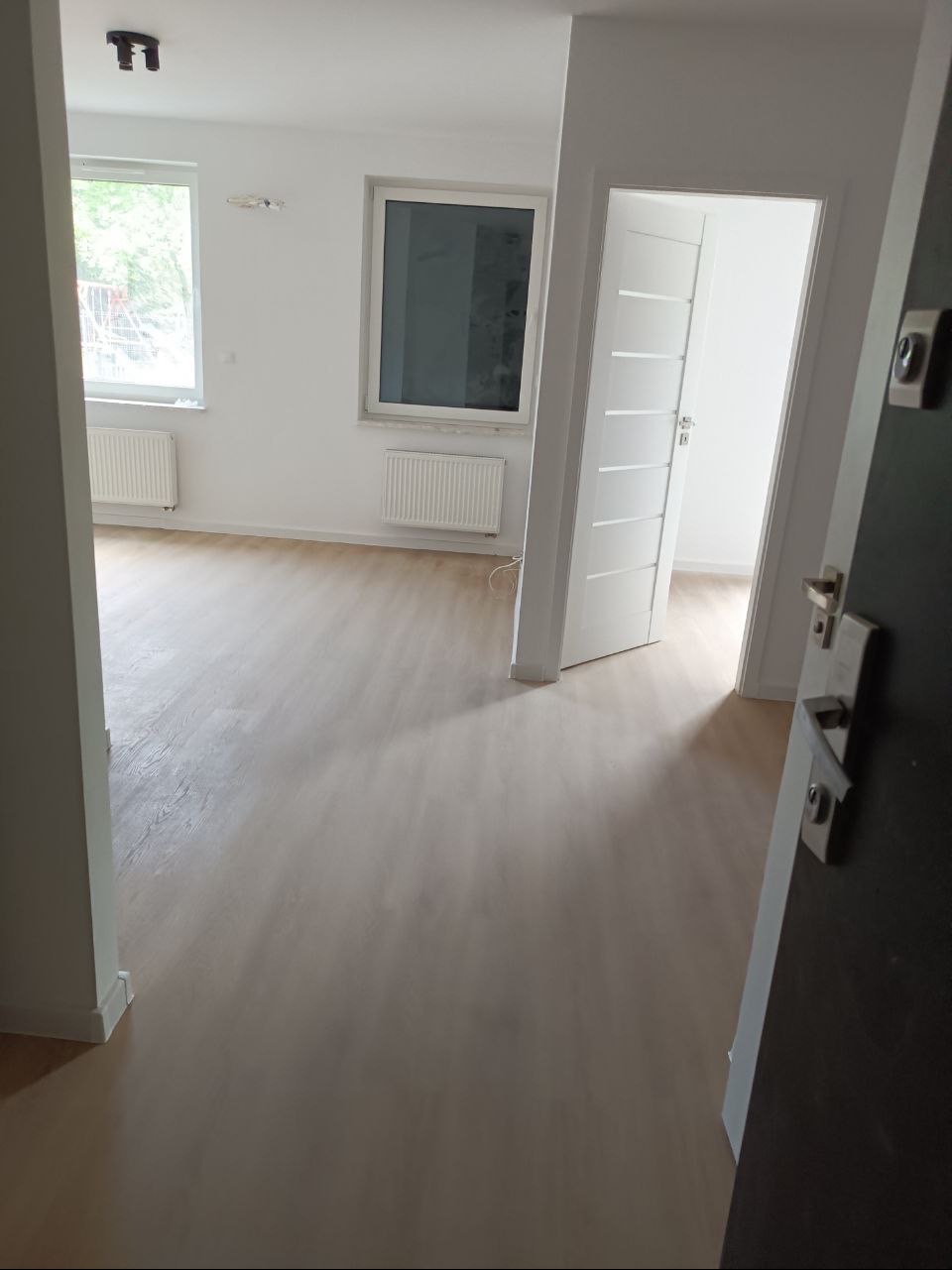 Widok zza uchylonych drzwi wejściowych na świeżo wykończone, jasne wnętrze mieszkania z nową podłogą, białymi ścianami, grzejnikami i drzwiami wewnętrznymi.