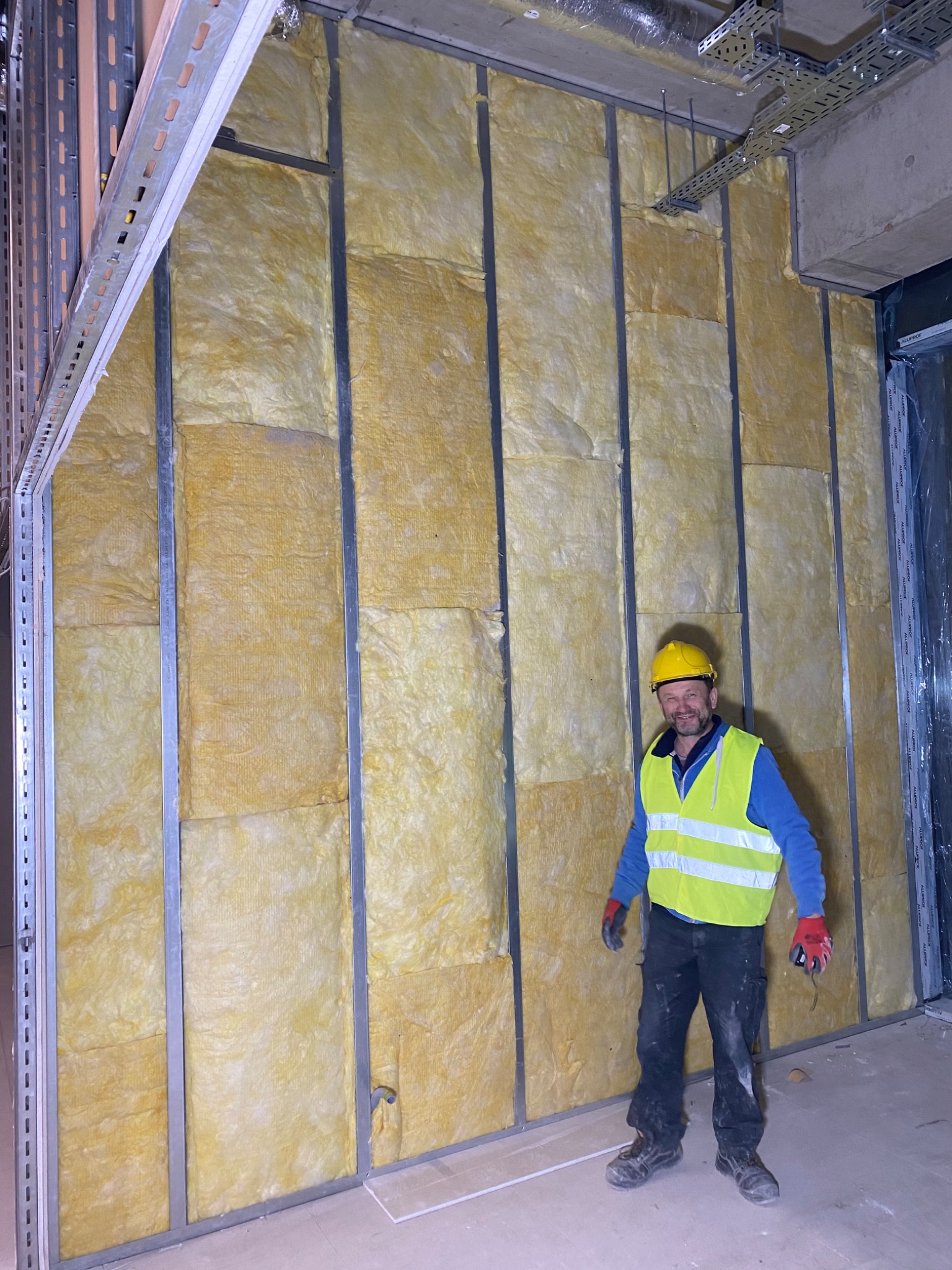Pracownik w żółtym kasku i kamizelce odblaskowej stoi na tle ściany z metalowym stelażem wypełnionym wełną mineralną, widoczne instalacje pod sufitem.