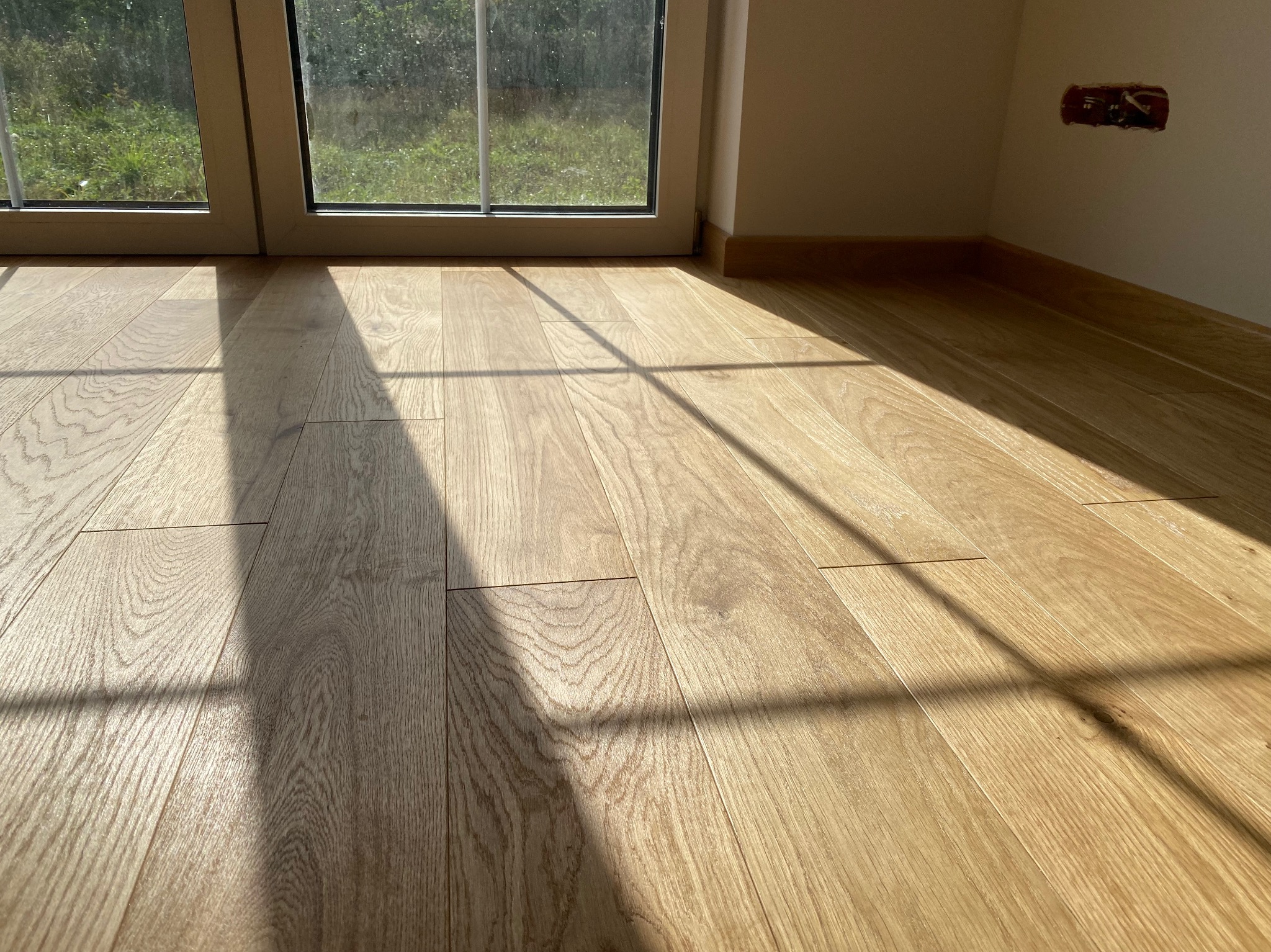 Jasna, drewniana podłoga ułożona w nowym mieszkaniu w Grabicy, oświetlona promieniami słońca wpadającymi przez okno, widoczny fragment ściany z puszką elektryczną.