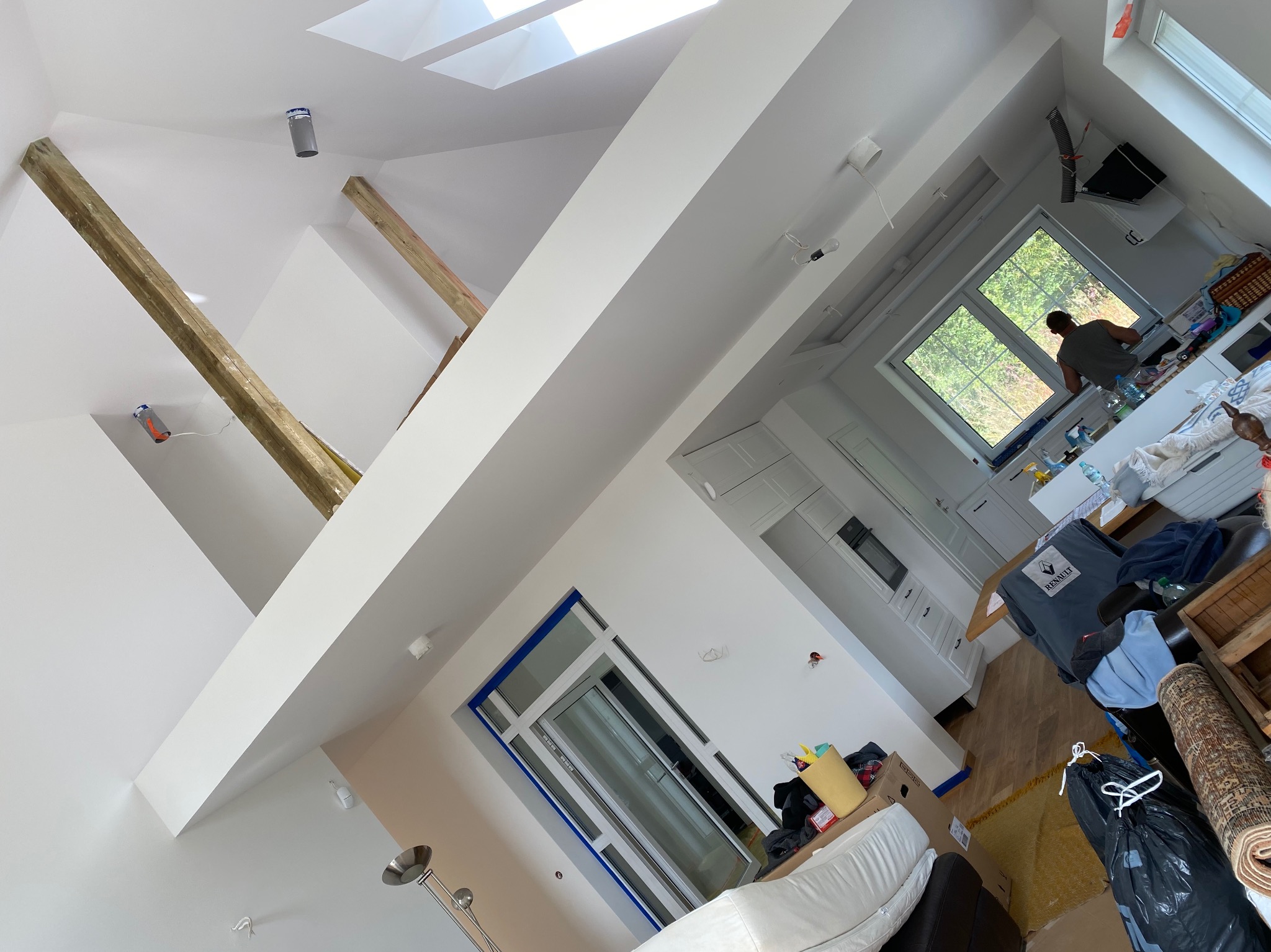 Wnętrze poddasza w trakcie prac wykończeniowych: widoczne drewniane belki konstrukcyjne, okno dachowe, białe ściany, okno balkonowe zabezpieczone taśmą malarską, oraz elementy wyposażenia w trakcie...