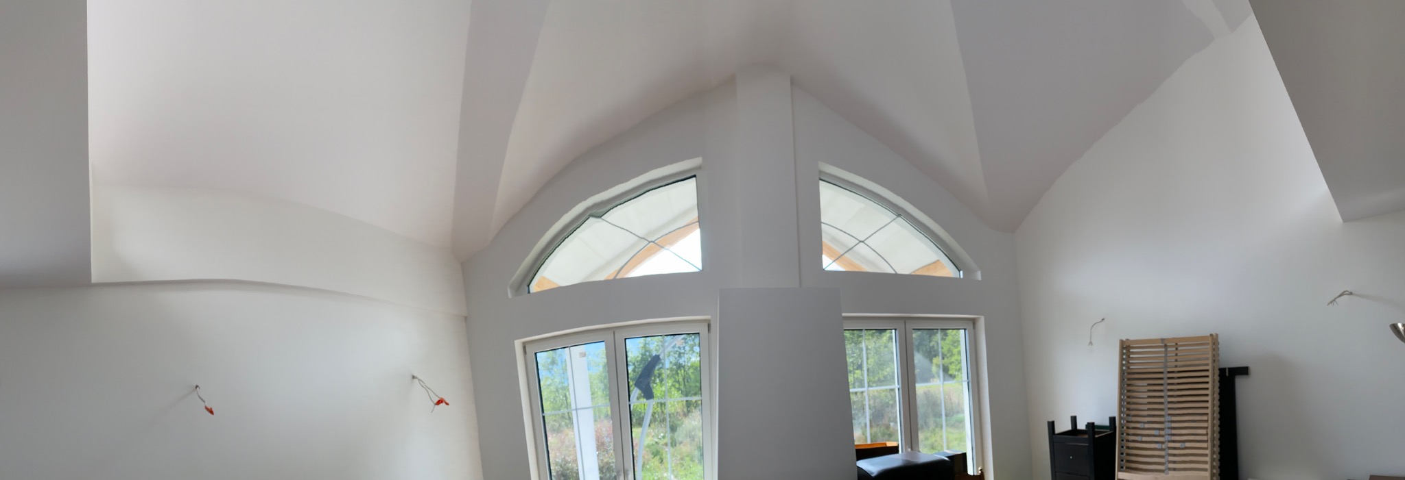 Wnętrze pokoju z wysokim sufitem i dwoma oknami łukowymi, ściany pomalowane na biało, widoczne wystające przewody elektryczne i drewniane elementy mebli w tle.