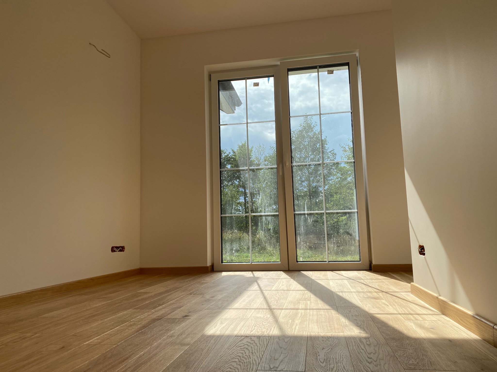 Jasne, puste pomieszczenie z drewnianą podłogą, dużym oknem z widokiem na zieleń i wystającymi kablami ze ściany, wpadające promienie słoneczne.