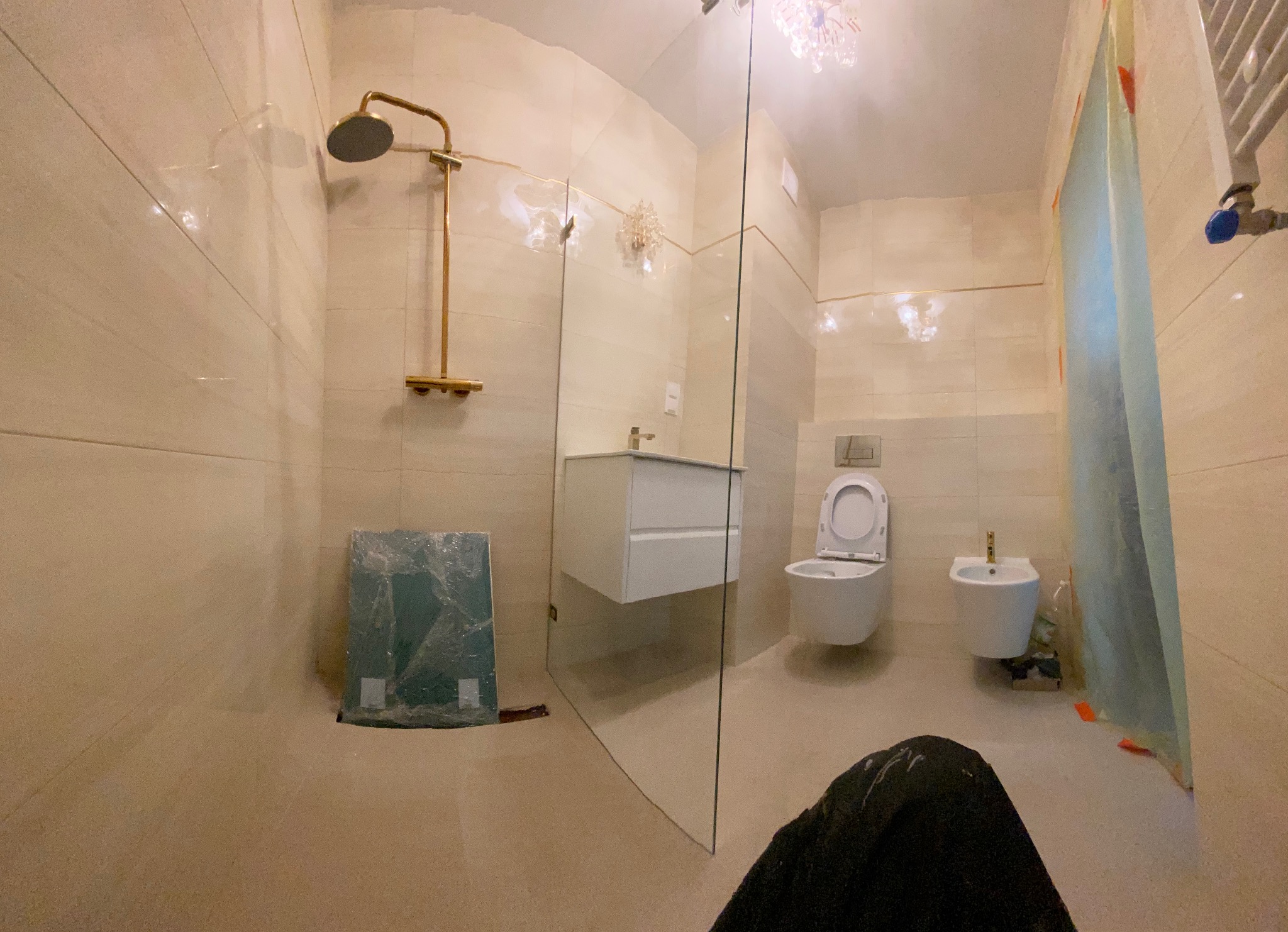 Jasna łazienka po remoncie z kabiną prysznicową typu walk-in, złotą armaturą i podwieszaną toaletą oraz bidetem, widoczne zabezpieczenia folią malarską i kartony z płytkami.