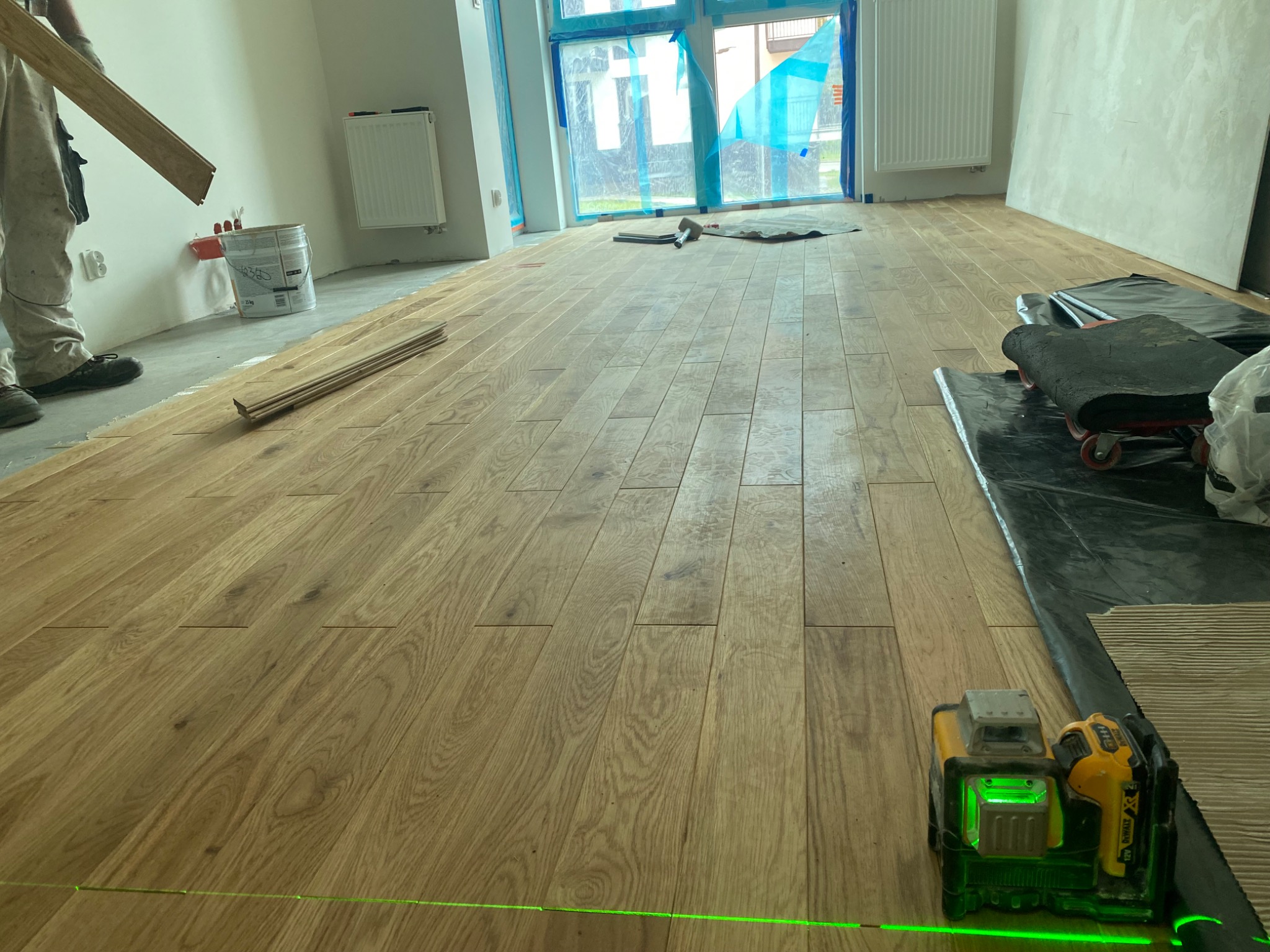 Układanie drewnianej podłogi w jasnym pomieszczeniu, widoczny laser krzyżowy, częściowo ułożone panele i narzędzia pracy.