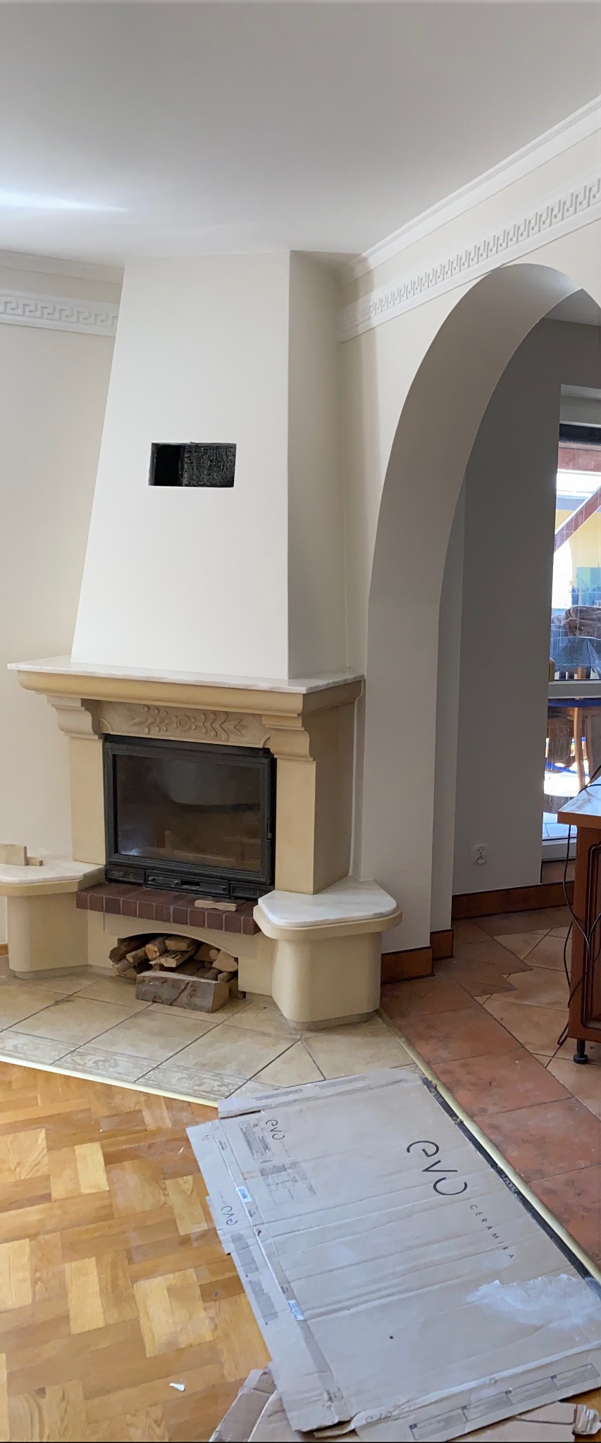 Klasyczny kominek z kamiennym obramowaniem i miejscem na drewno, widoczne kartony z ceramiką na podłodze, fragment łuku w tle, dekoracyjny gzyms przy suficie.
