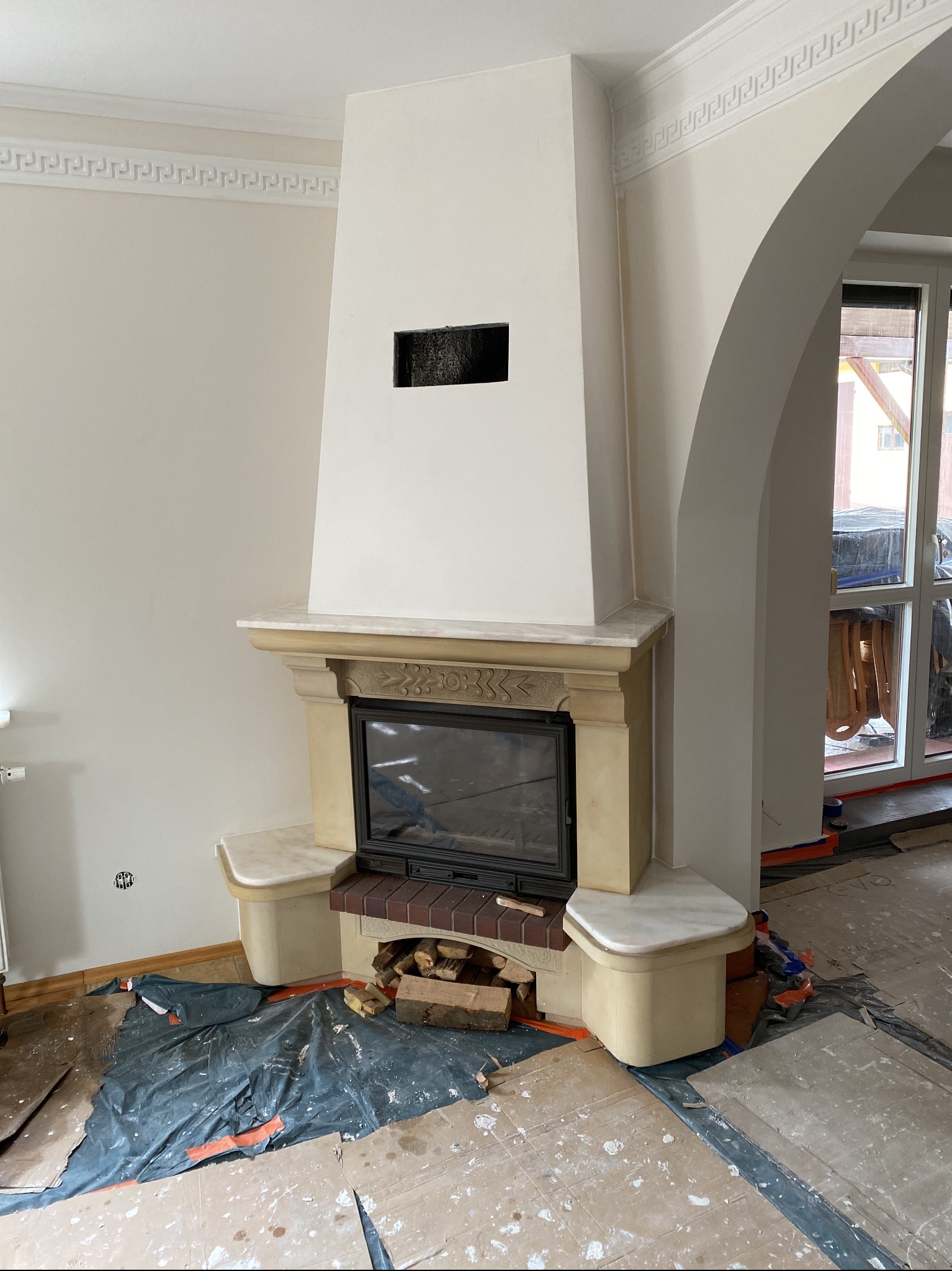 Wnętrze z kominkiem w trakcie prac wykończeniowych; zabezpieczona podłoga, widoczne dekoracyjne listwy przysufitowe i łukowe przejście.