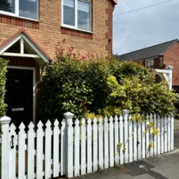 Białe, drewniane ogrodzenie typu sztachety przed ceglanym domem, z zielenią w tle oraz czarnymi drzwiami wejściowymi z numerem 55.